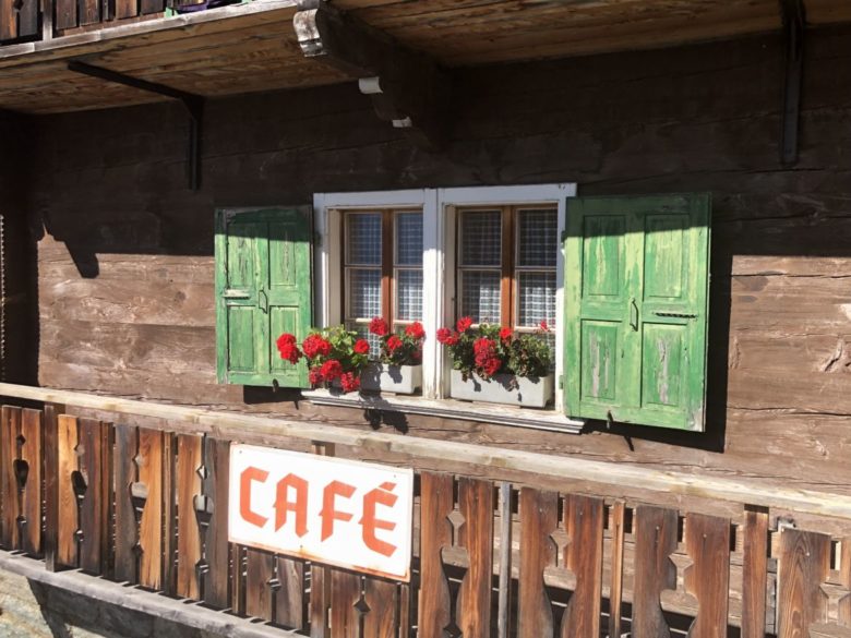 Photo: Un balcon en bois, une pancarte "Café" en rouge sur fond blanc; une fenêtre avec des géranium et des volets verts.